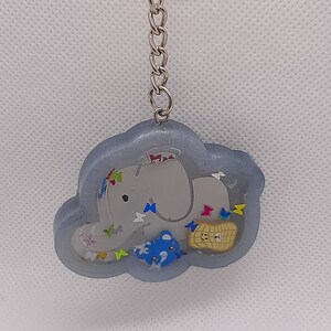 Puede incluir: Un llavero de resina gris en forma de elefante con un elefante azul, una cesta marrón y mariposas de colores en su interior. El llavero tiene un anillo y una cadena de metal plateado.