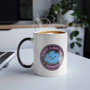 Puede incluir: Una taza de cerámica blanca con asa negra, con un diseño "ALWAYS SLEEPY CLUB MEMBER". El diseño incluye una nube azul sonriente en un marco circular rosa y negro. Sale vapor del café dentro de la taza.