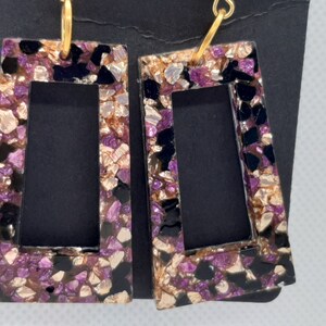 May include: A pair of gold hoop earrings with a black, purple, and gold rectangular frame. The frame is filled with small pieces of glitter.