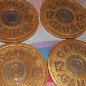 May include: Four gold-colored coasters with the text "GAUGE 12" in a circular design. The coasters have a metallic sheen and a central circular detail. The overall design resembles a shotgun shell.