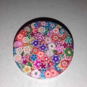 May include: A round, white phone grip with a colorful floral design. The grip is covered in small, multi-colored flowers.