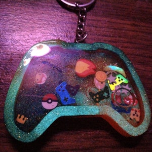 Op de afbeelding: Een groene glitterhars sleutelhanger in de vorm van een videogamecontroller. De controller is gevuld met kleurrijke bedels, waaronder een Pokémon-personage, een ster en een vuursymbool.