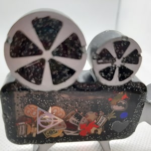 May include: A gray film camera figurine with white film reels. The camera's body is filled with small, colorful movie-themed charms, including a teddy bear, a ticket, and a Batman logo. The figurine has three small legs.