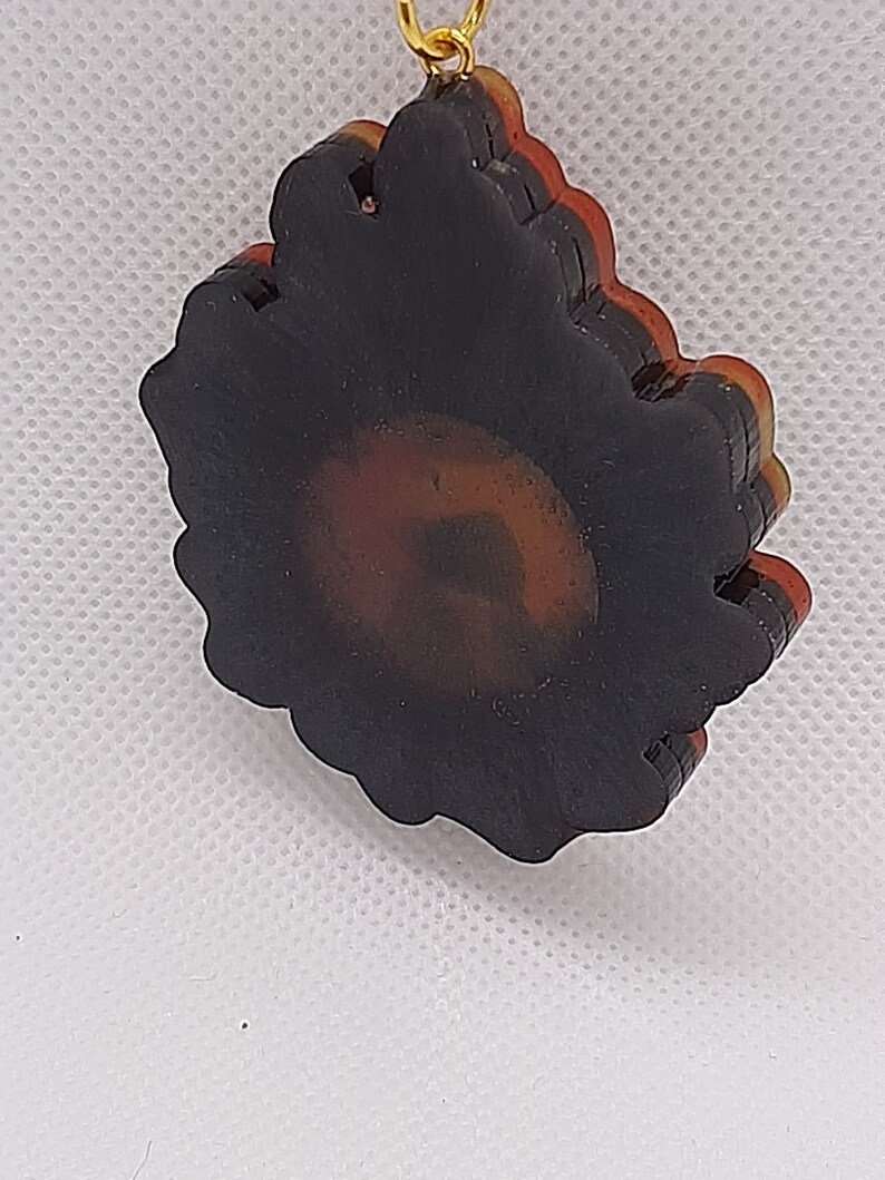 May include: A layered black, red, and gold resin sunflower pendant with a gold chain.