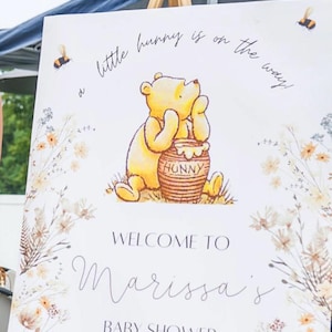 K&ouml;nnte beinhalten: Ein wei&szlig;es Schild mit einer Winnie-the-Pooh-Illustration und floralen Akzenten. Das Schild lautet "little bunny is on the way!" und "Welcome to Marissa's Baby Shower July 20th 2024".