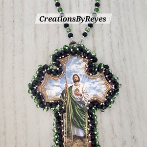May include: A wooden cross with a beaded border of black and green crystals. The cross features a printed image of Saint Jude Thaddeus, a Catholic saint known for miracles. The text "Amigo de Jesus!" is printed at the bottom of the image.