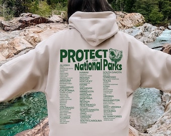 Op de afbeelding: Een beige hoodie met de tekst "Bescherm onze nationale parken" in groene letters. De hoodie heeft een lijst met alle nationale parken in de Verenigde Staten, inclusief Alaska en Hawaï.