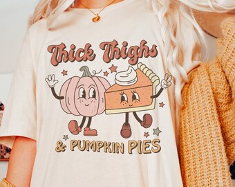 Puede incluir: Una camiseta blanca con un gráfico de una calabaza y una rebanada de pastel de calabaza con el texto "Thick Thighs & Pumpkin Pies".