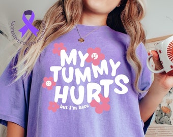 Puede incluir: Una camiseta de color lila claro con el texto "MY TUMMY HURTS but I'm here" en blanco, rodeado de flores rosas. La camiseta también tiene una cinta morada con las palabras "Ulcerative Colitis Awareness". Una persona sostiene una taza con un diseño de sol.