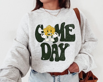 Op de afbeelding: Een lichtgrijze sweatshirt met de woorden "GAME DAY" in donkergroen, met een cartoon kaaswig karakter. De kaaswig heeft een lachend gezicht, witte handschoenen en witte schoenen. De sweatshirt heeft lange mouwen.