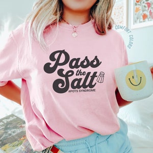 May include: A pink t-shirt with the text "Pass the Salt" in a retro font. The text is in black and includes a small salt shaker icon. Below the text is the hashtag "#POTS SYNDROME".