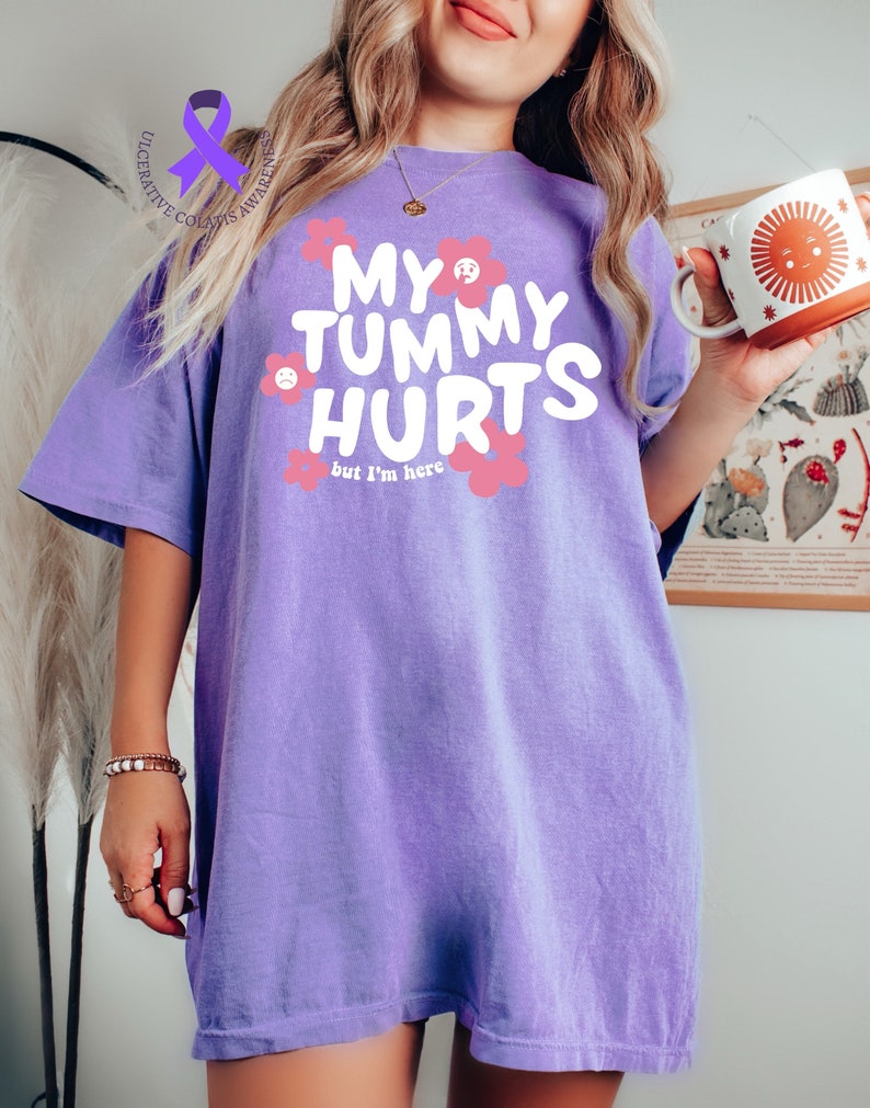 May include: A light purple t-shirt with the text "MY TUMMY HURTS but I'm here" in white, surrounded by pink flowers. The shirt also has a purple ribbon with the words "Ulcerative Colitis Awareness". A person is holding a mug with a sun design.