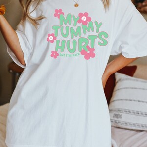 May include: White t-shirt with the text "MY TUMMY HURTS" in green bubble letters, accented with pink flowers. The phrase "but I'm here" is below the main text. The shirt has short sleeves and a relaxed fit.