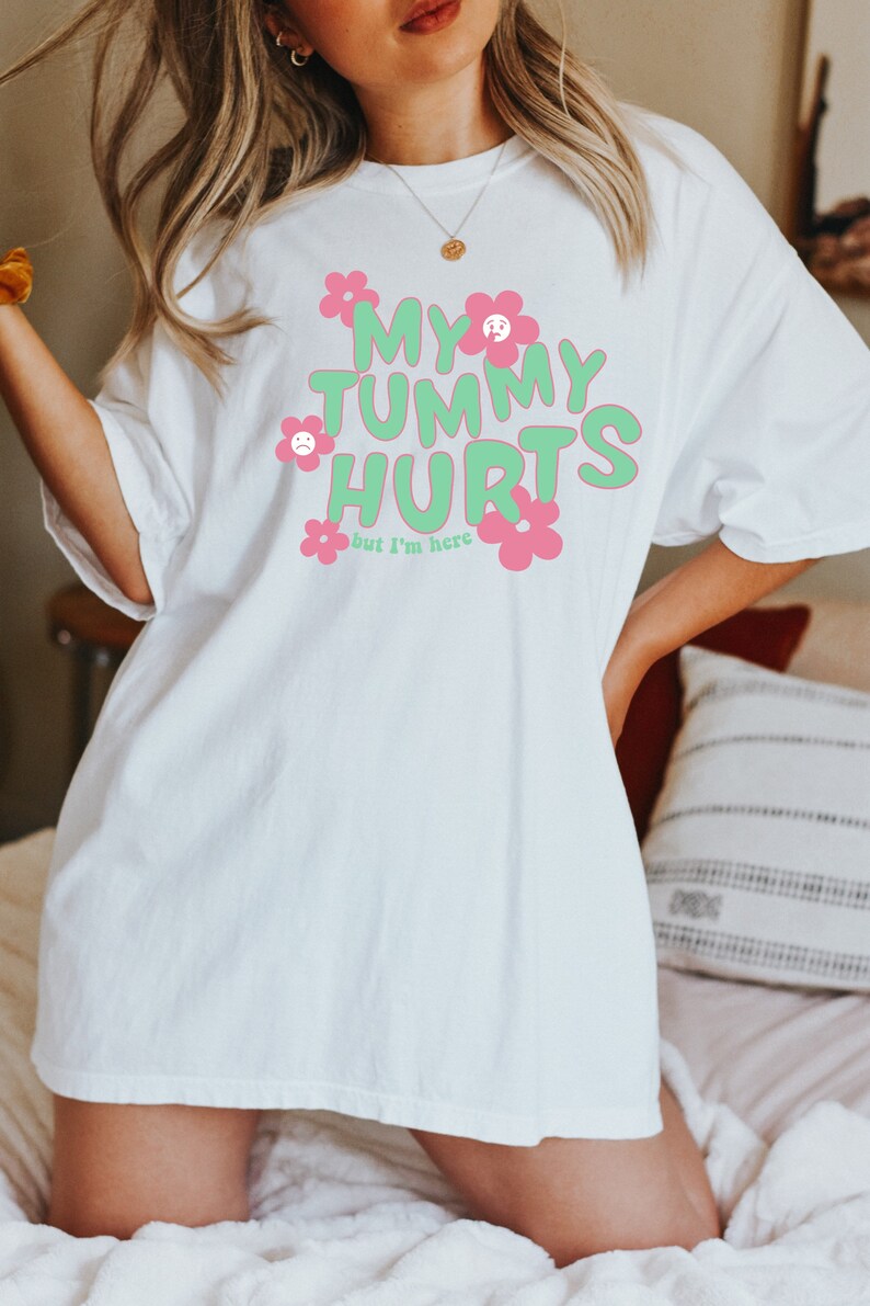 May include: White t-shirt with the text "MY TUMMY HURTS" in green bubble letters, accented with pink flowers. The phrase "but I'm here" is below the main text. The shirt has short sleeves and a relaxed fit.