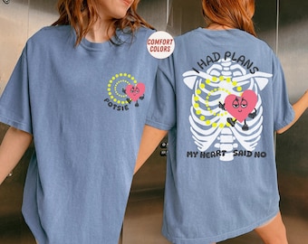 Op de afbeelding: Blauw t-shirt met een afbeelding op de voor- en achterkant. De voorkant heeft een roze hartkarakter met het woord "Potsie" en een geel spiraalontwerp. De achterkant heeft een skeletontwerp met een roze hart en de tekst "I had plans, my heart said no."