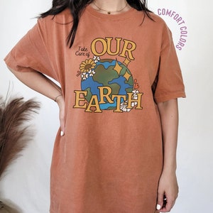 Puede incluir: Una camiseta de color &oacute;xido con un gr&aacute;fico de una tierra de dibujos animados con las palabras "Take Care of Our Earth" impresas en ella. El gr&aacute;fico est&aacute; rodeado de flores y estrellas.