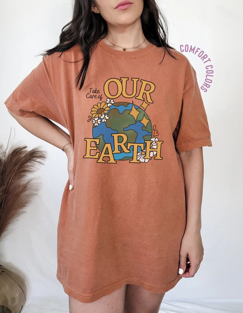 Puede incluir: Una camiseta de color &oacute;xido con un gr&aacute;fico de una tierra de dibujos animados con las palabras "Take Care of Our Earth" impresas en ella. El gr&aacute;fico est&aacute; rodeado de flores y estrellas.