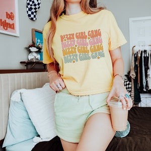 Op de afbeelding: Een geel gecropped t-shirt met de tekst "Dizzy Girl Gang" die vier keer wordt herhaald in een retro lettertype. Elke regel tekst is in een andere kleur, met roze, oranje en groen. De tekst is versierd met kleine gele bloemen.