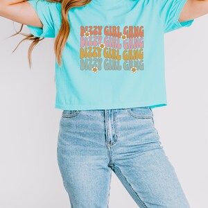 Op de afbeelding: Een turquoiseblauw gecropped t-shirt met de tekst "Dizzy Girl Gang" die vier keer wordt herhaald in een retro lettertype met margriet accenten.