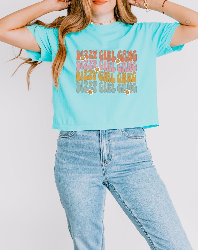 Op de afbeelding: Een turquoiseblauw gecropped t-shirt met de tekst "Dizzy Girl Gang" die vier keer wordt herhaald in een retro lettertype met margriet accenten.