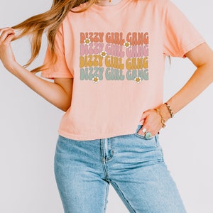 Op de afbeelding: Een perzikkleurig T-shirt met de tekst "Dizzy Girl Gang" die vier keer wordt herhaald in een retro-lettertype. Elke regel tekst is in een andere kleur, met een klein madeliefje-grafisch ontwerp naast elke regel tekst.