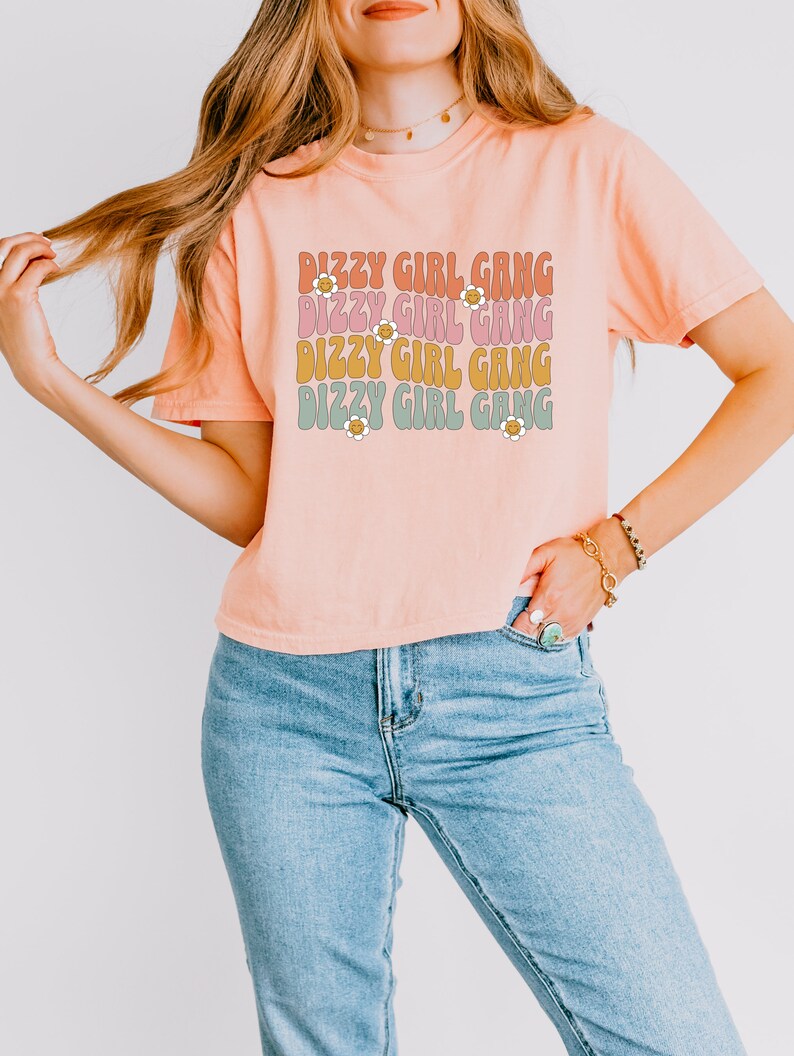 Op de afbeelding: Een perzikkleurig T-shirt met de tekst "Dizzy Girl Gang" die vier keer wordt herhaald in een retro-lettertype. Elke regel tekst is in een andere kleur, met een klein madeliefje-grafisch ontwerp naast elke regel tekst.
