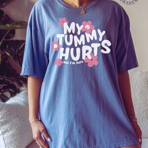 May include: Blue t-shirt with the text "MY TUMMY HURTS" in white, surrounded by pink flowers. The phrase "but I'm here" is below the main text. The shirt is part of IBS awareness.