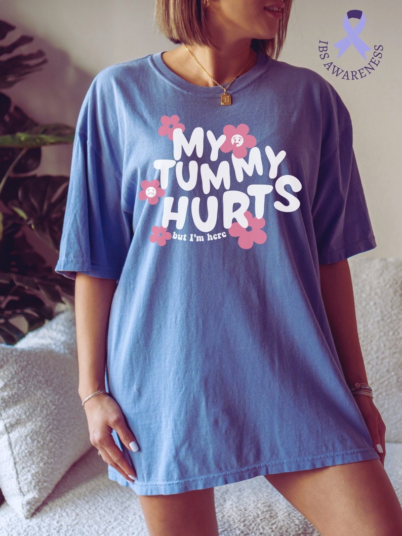 May include: Blue t-shirt with the text "MY TUMMY HURTS" in white, surrounded by pink flowers. The phrase "but I'm here" is below the main text. The shirt is part of IBS awareness.