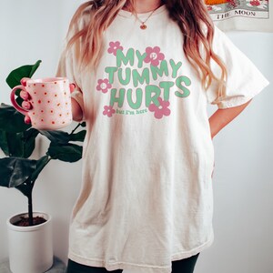 May include: Cream-colored t-shirt with the text "MY TUMMY HURTS but I'm here" in green and pink, with flower graphics. The shirt is worn by a person holding a pink mug with orange polka dots.