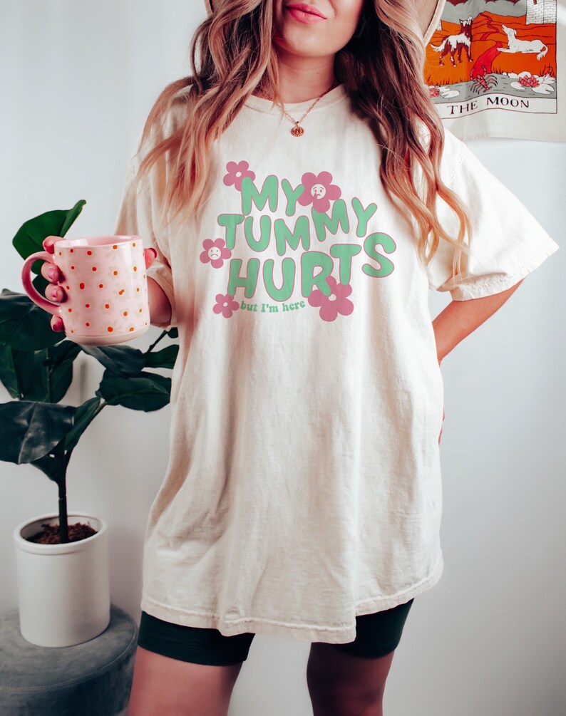 May include: Cream-colored t-shirt with the text "MY TUMMY HURTS but I'm here" in green and pink, with flower graphics. The shirt is worn by a person holding a pink mug with orange polka dots.