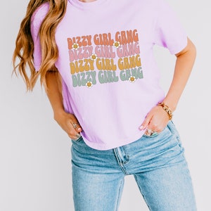 Op de afbeelding: Een lichtpaarse T-shirt met de tekst "Dizzy Girl Gang" die vier keer wordt herhaald in een retro-lettertype met een madeliefje-afbeelding. De tekst is in tinten roze, oranje en groen.