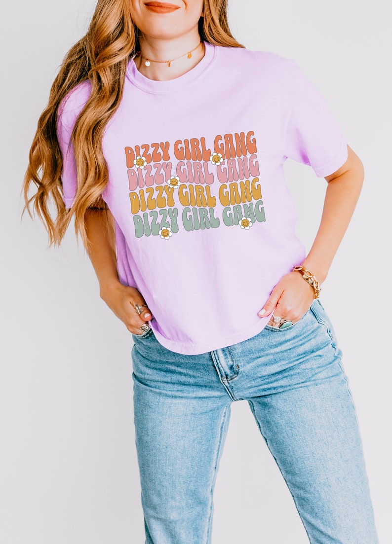 Op de afbeelding: Een lichtpaarse T-shirt met de tekst "Dizzy Girl Gang" die vier keer wordt herhaald in een retro-lettertype met een madeliefje-afbeelding. De tekst is in tinten roze, oranje en groen.