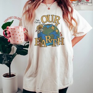 Puede incluir: Una camiseta blanca con un gr&aacute;fico de una tierra azul y verde con flores y el texto "Take Care of Our Earth".