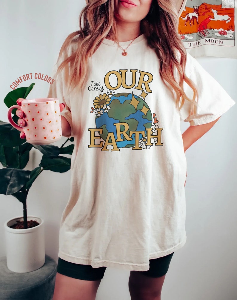 Puede incluir: Una camiseta blanca con un gr&aacute;fico de una tierra azul y verde con flores y el texto "Take Care of Our Earth".