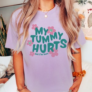 May include: A light purple t-shirt with the text "MY TUMMY HURTS but I'm here" in teal and pink. The shirt features a crew neck and short sleeves. The graphic includes flower designs with sad faces.