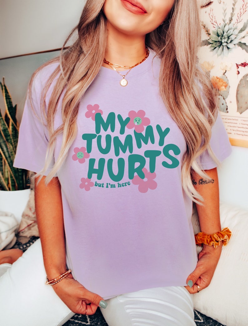 May include: A light purple t-shirt with the text "MY TUMMY HURTS but I'm here" in teal and pink. The shirt features a crew neck and short sleeves. The graphic includes flower designs with sad faces.