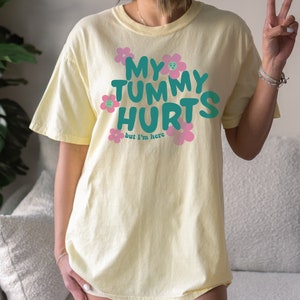 May include: A light yellow t-shirt with the text "MY TUMMY HURTS but I'm here" in teal and pink. The text is surrounded by pink flowers with sad faces. The shirt is a casual, oversized fit.
