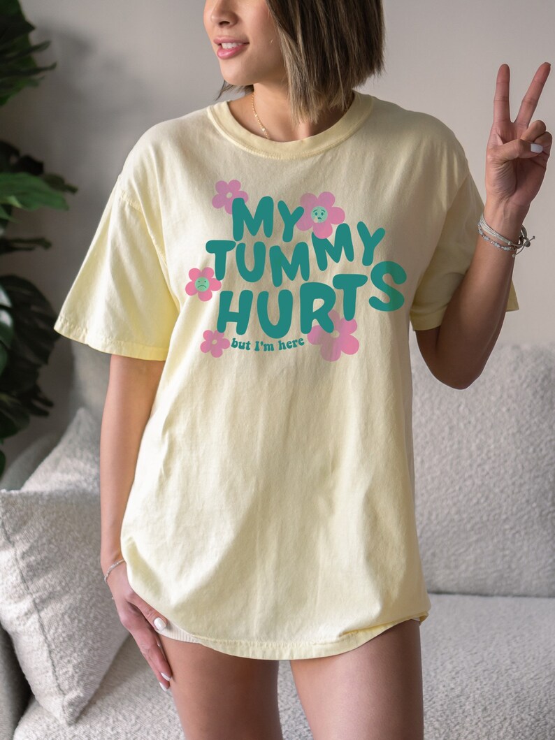 May include: A light yellow t-shirt with the text "MY TUMMY HURTS but I'm here" in teal and pink. The text is surrounded by pink flowers with sad faces. The shirt is a casual, oversized fit.