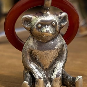 May include: A silver monkey pendant with a red circular accent. The monkey is sitting with its legs extended. The pendant is made of a shiny metal and has a detailed design. The red accent is a smooth, polished ring.