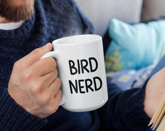 May include: A person sitting on a couch holding a white mug with the text "BIRD NERD" in black lettering. The person is reading a book.