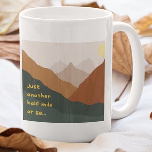 May include: White ceramic mug with a graphic of a mountain range with a sun in the sky. The text on the mug reads "Just another half mile or so..."