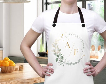May include: White apron with black straps featuring a gold and green floral wreath design. The wreath surrounds the initials "A/F" and the name "ASHLEY FISHER". The apron is worn in a kitchen setting.