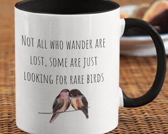 May include: White ceramic mug with a black rim and handle. The mug has a quote that reads "Not all who wander are lost, some are just looking for rare birds." The quote is printed in black text. The mug features a watercolor illustration of two birds perched on a branch.