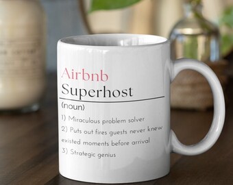 May include: White ceramic mug with the text "Airbnb Superhost" in black and pink. The mug has a definition of "Superhost" with three points. The mug is on a dark wooden surface.