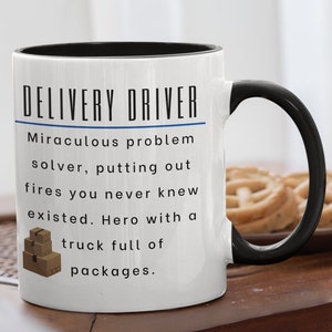May include: White ceramic mug with a black handle and rim. The mug features the text "DELIVERY DRIVER" above a description of a delivery driver's role. A stack of brown boxes is also shown on the mug.
