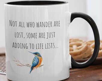 May include: White ceramic mug with a black rim and handle. The mug features a watercolor illustration of a blue and orange bird perched on a branch. The text on the mug reads "Not all who wander are lost, some are just adding to life lists..."