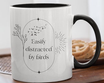 May include: White ceramic mug with a black rim and handle. The mug has a black and white design with the text "Easily distracted by birds" and a stylized image of birds flying and a sun.