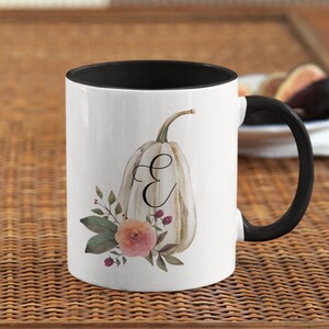 May include: White ceramic mug with a black rim and handle. The mug features a white pumpkin with a floral design and the letter "E" on the front.