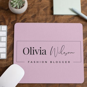 May include: A pink mouse pad with a black border and the text "Olivia Wilson FASHION BLOGGER" in black.