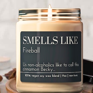 Peut inclure: Un bocal en verre pour bougie avec un couvercle doré et une étiquette noire sur laquelle est inscrit "SMELLS LIKE Fireball Us non-alcoholics like to call this cinnamon Becky... 100% vegan soy wax blend | 9oz | non-toxic". La bougie est allumée et la cire est fondue.